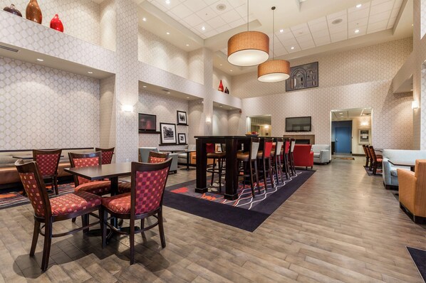 A spacious hotel lobby with a high ceiling, multiple seating areas including a long dining table and booths, and a television mounted on the wall.