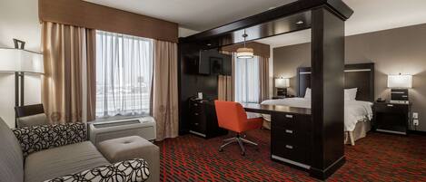 Habitación de hotel con sofá, escritorio, silla, cama, televisor y ventana con cortinas.