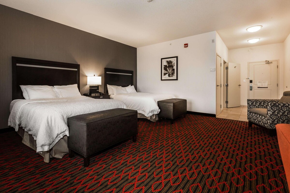 Habitación de hotel con dos camas, un ventanal amplio, alfombra roja con estampado y una pequeña zona de estar.