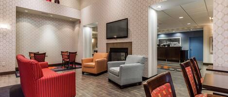 Un hall de hotel con zona de estar que incluye sofás rojos y grises, un televisor de pantalla plana montado en la pared y una chimenea.