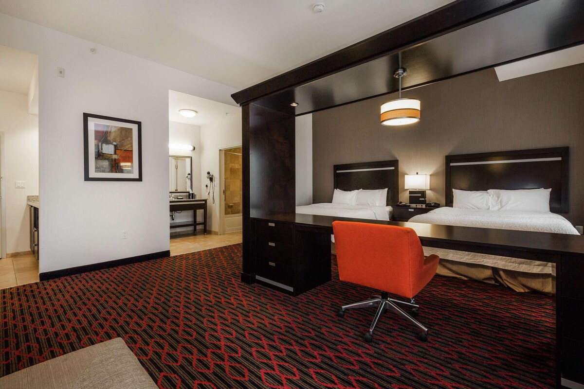 Habitación de hotel con una cama grande, un escritorio y una silla. Hay un espejo en la pared, un marco de fotos y una lámpara sobre el escritorio.