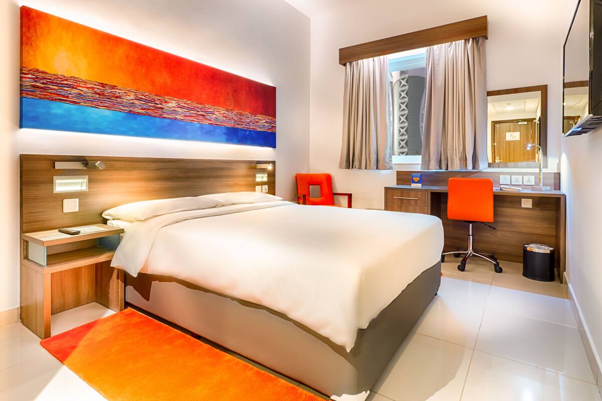 A modern hotel room with a large bed, a desk with a chair, a large window with curtains, and a painting on the wall.