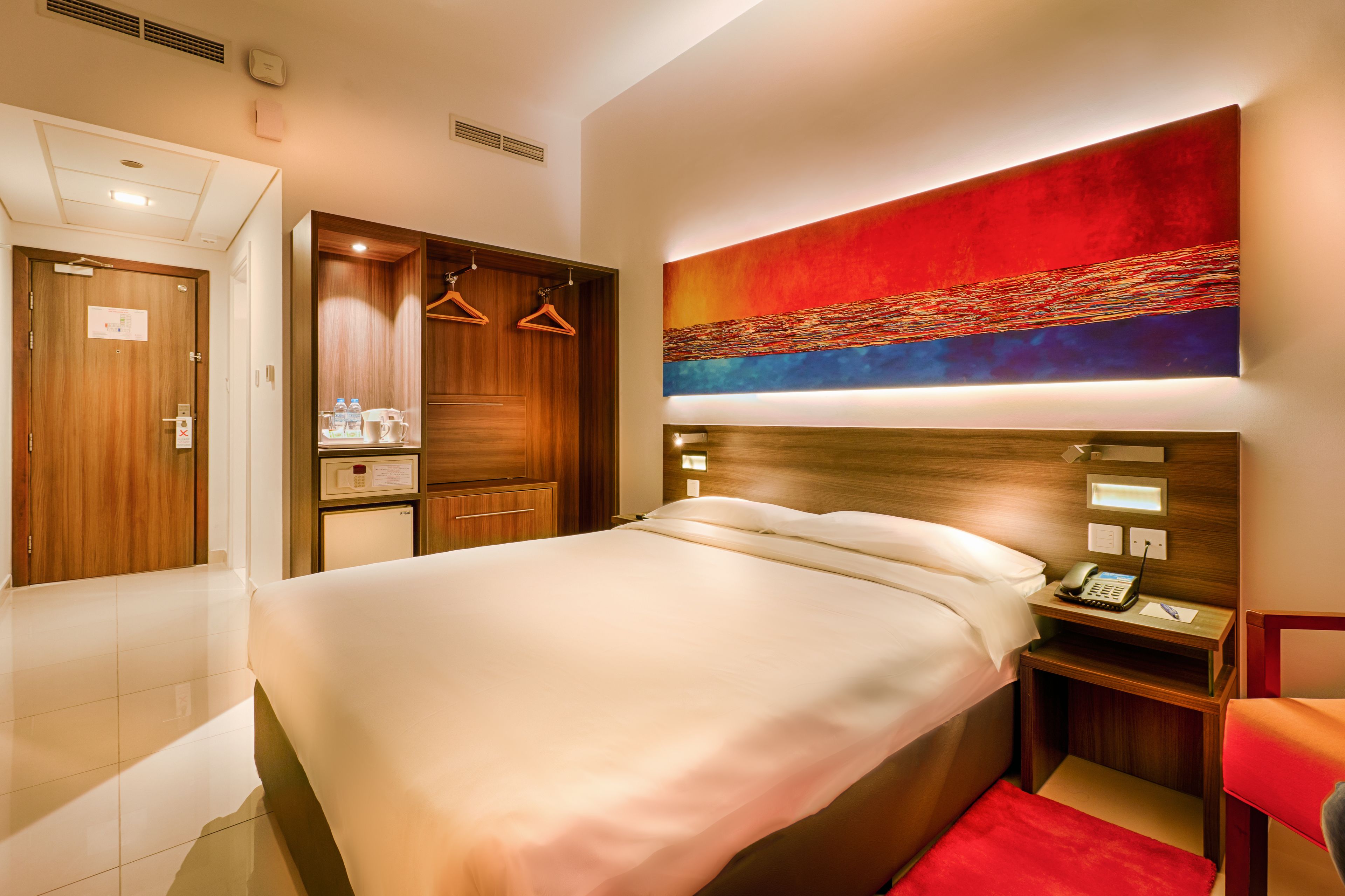 A hotel room with a large bed, a wooden wardrobe, a desk with a phone, and a red chair.