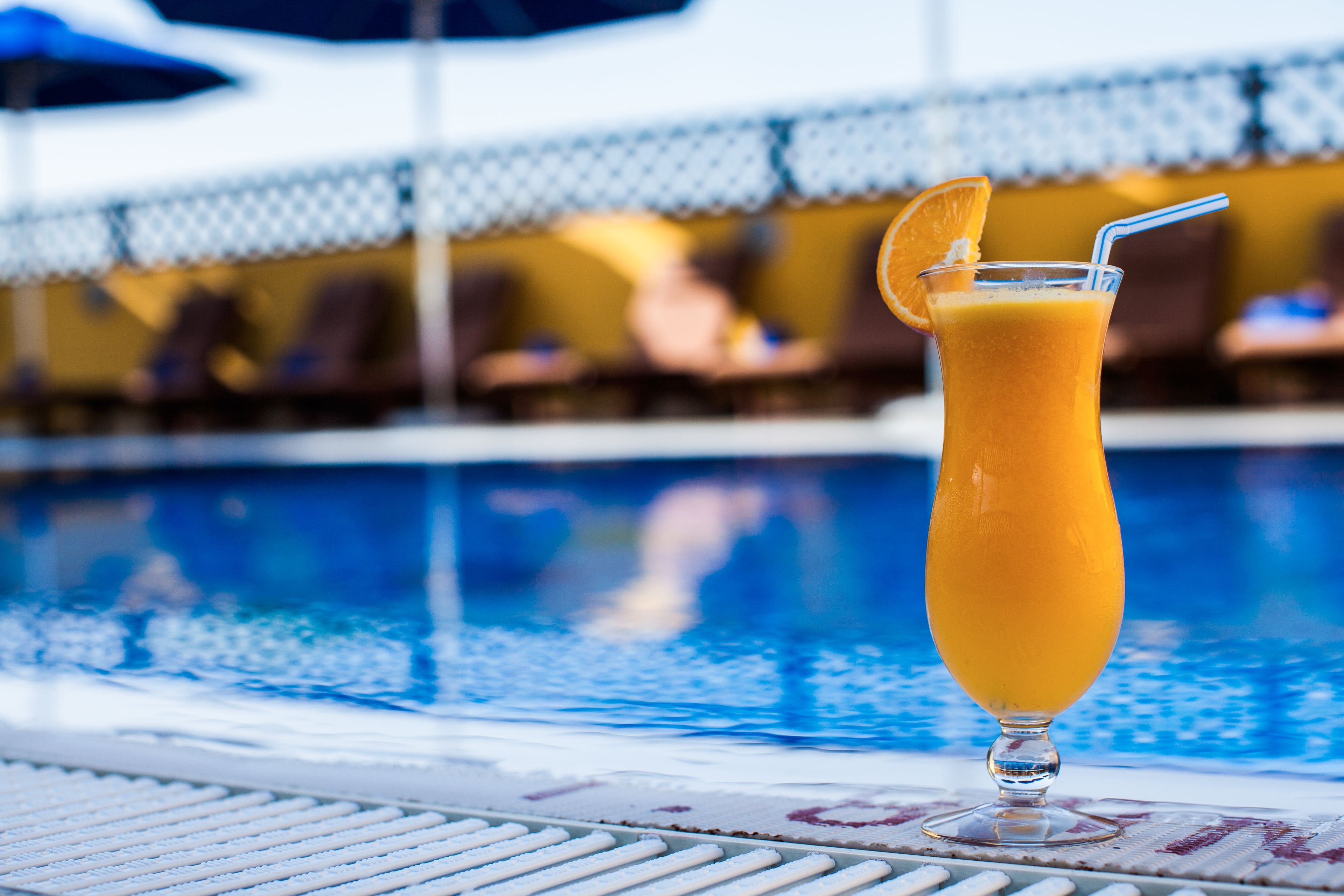 A glass of orange juice with a straw and a slice of orange on the rim, placed on a poolside table with a pool in the background.