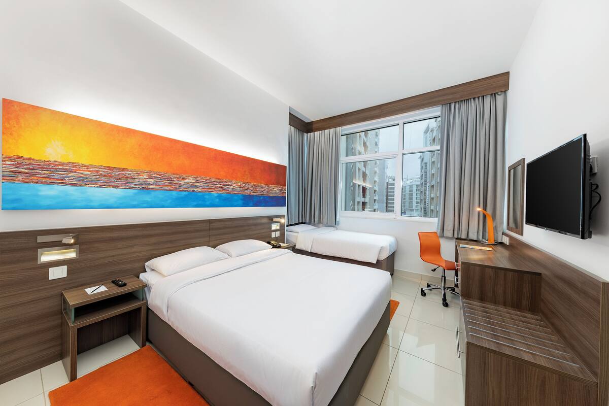 A hotel room with a large bed, a smaller bed, a desk with a chair, a TV, and a painting on the wall.