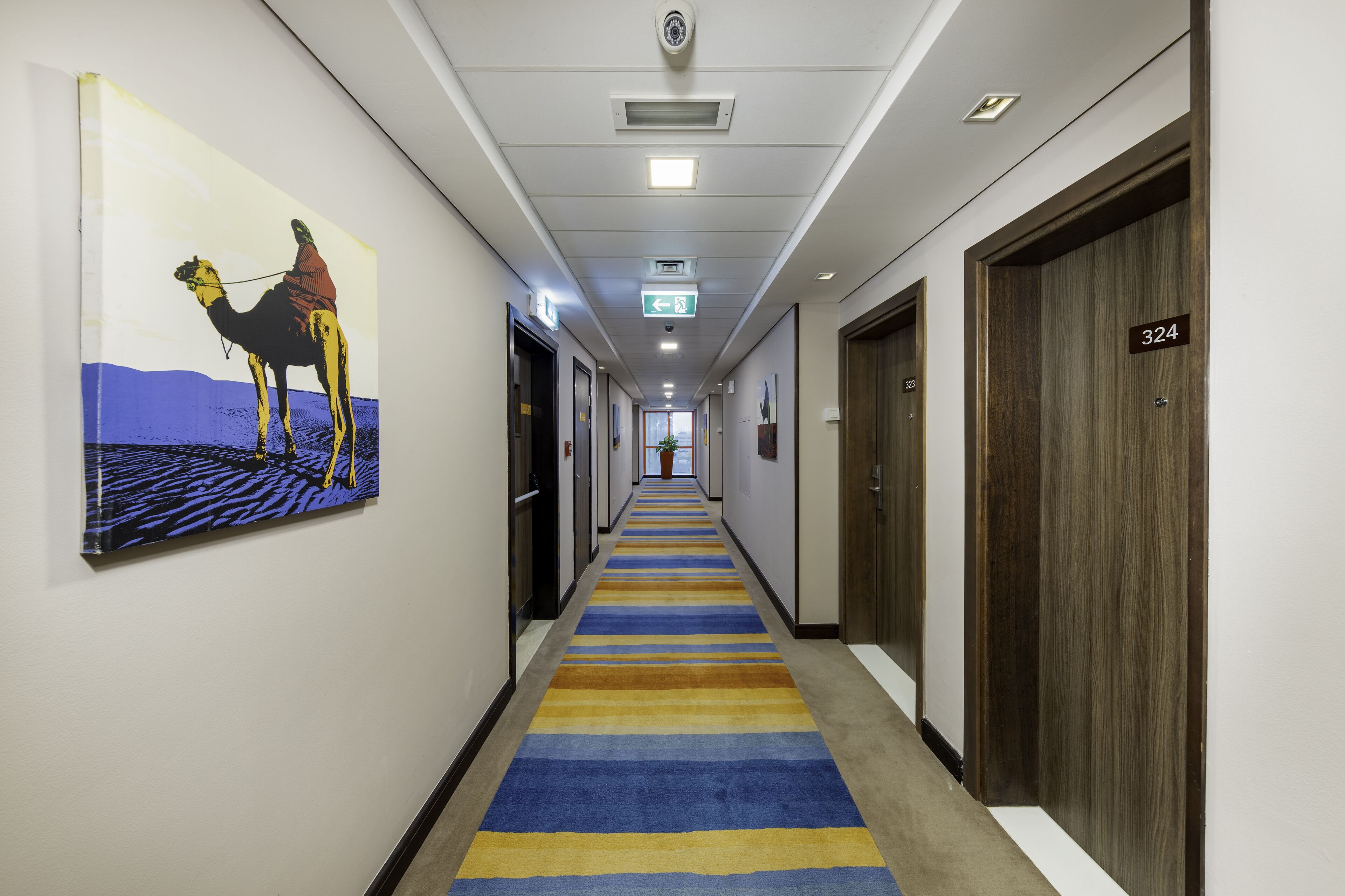 A hotel hallway with a striped carpet, a camel painting on the left wall, and doors numbered 324.