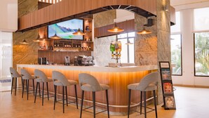 A bar area with a wooden counter, bar stools, a television, and a view of palm trees through the window.