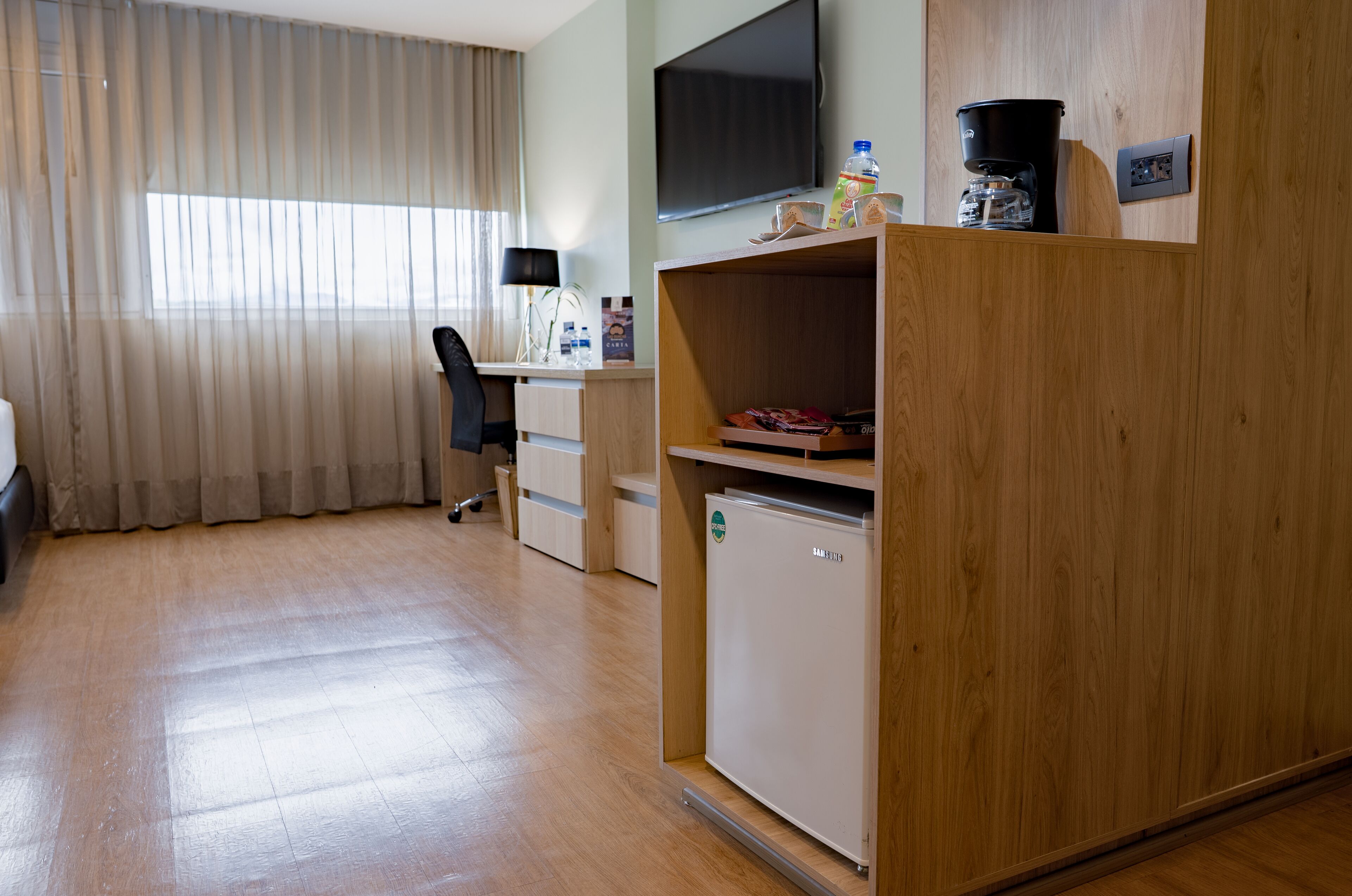A hotel room with a wooden desk, a chair, a small refrigerator, a television, and a window with curtains.