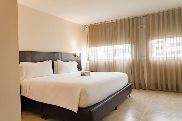 A large bed with a dark headboard, a bedside table with a lamp, and sheer curtains on a window.
