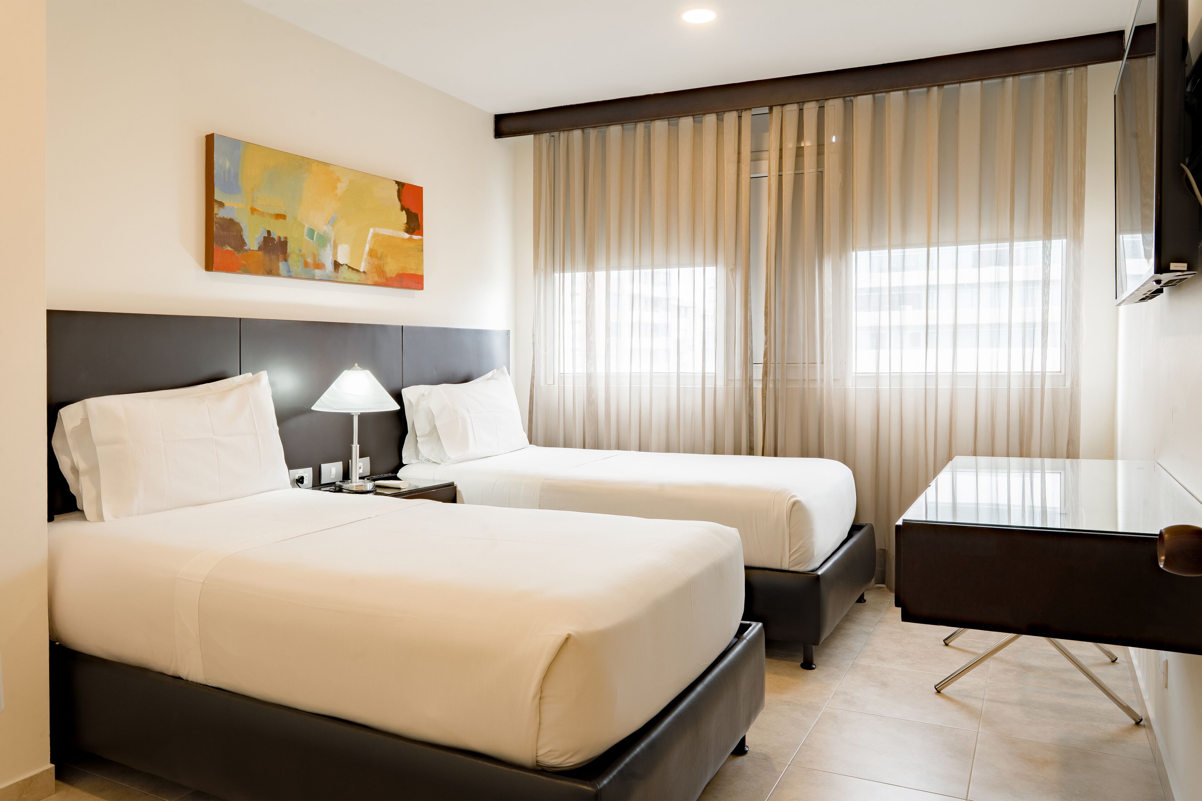 A hotel room with two beds, a desk, a lamp, a painting on the wall, and a large window with curtains.