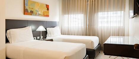 A hotel room with two beds, a desk, a lamp, a painting on the wall, and a large window with curtains.