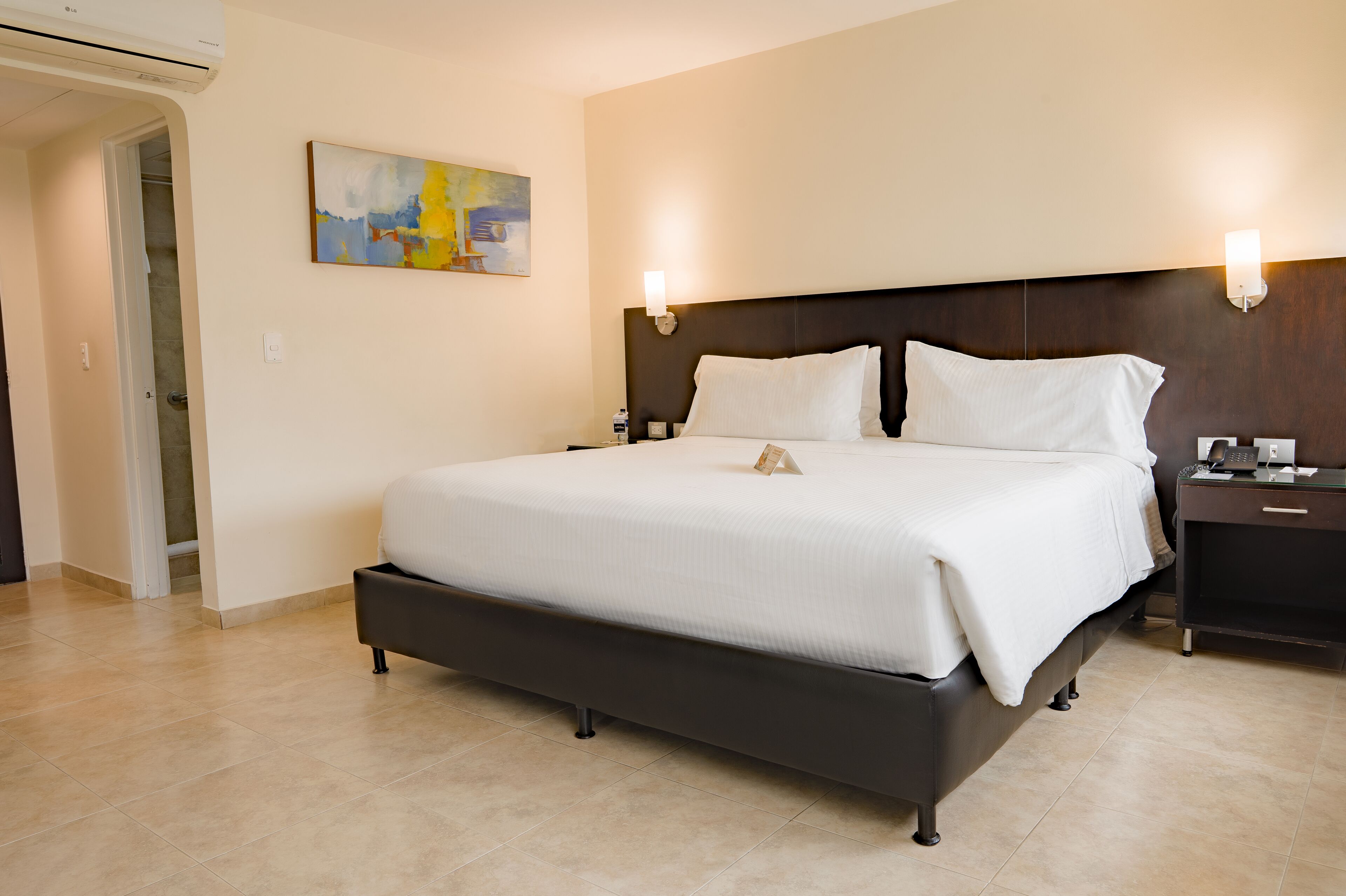 Ein Hotelzimmer mit einem großen Bett, Nachttischen, einem Wandbild und einer Klimaanlage.