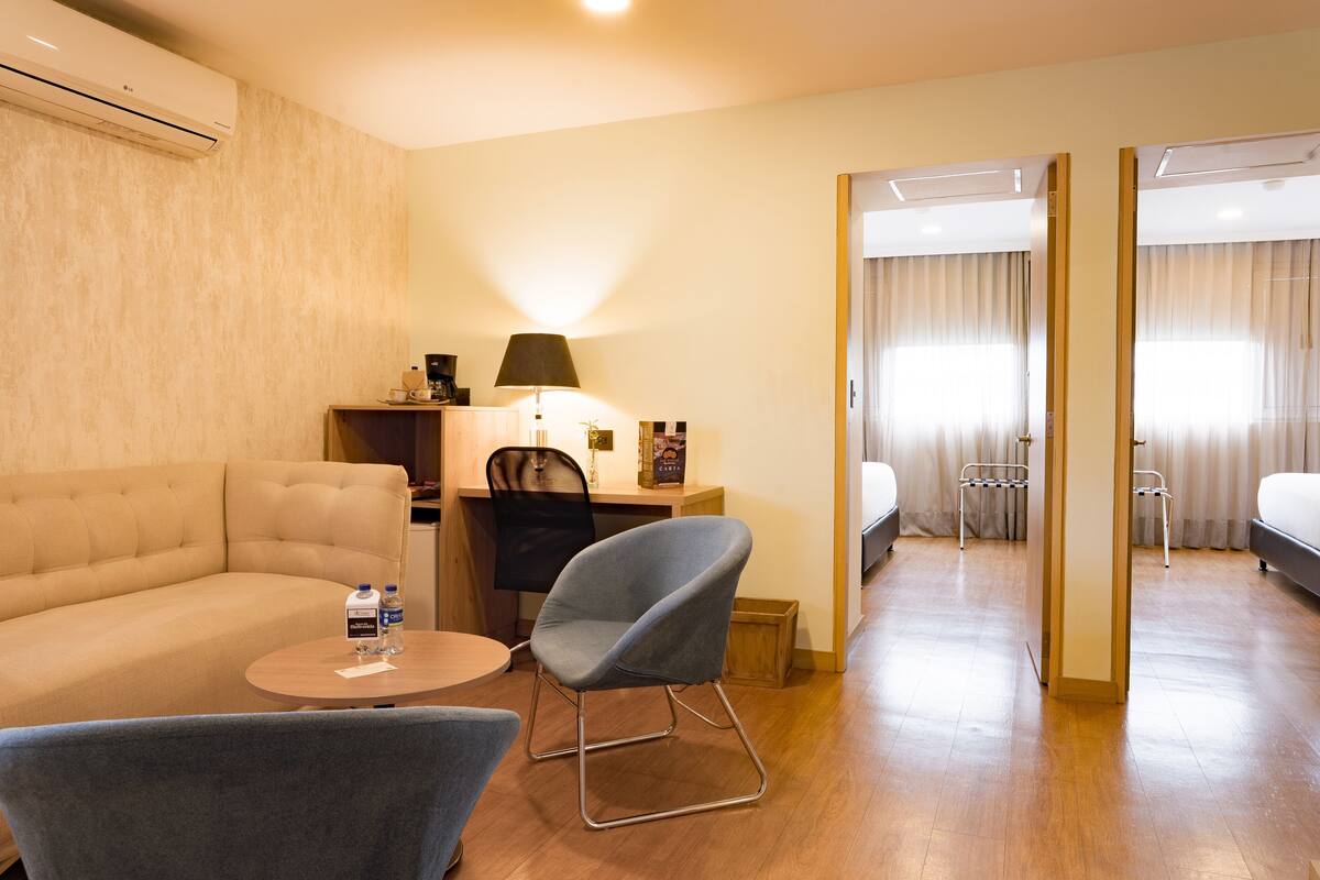 Ein Hotelzimmer mit Sofa, Schreibtisch mit Lampe, Stuhl, Bett und einem durchgegangene Tür erkennbaren Badezimmer.