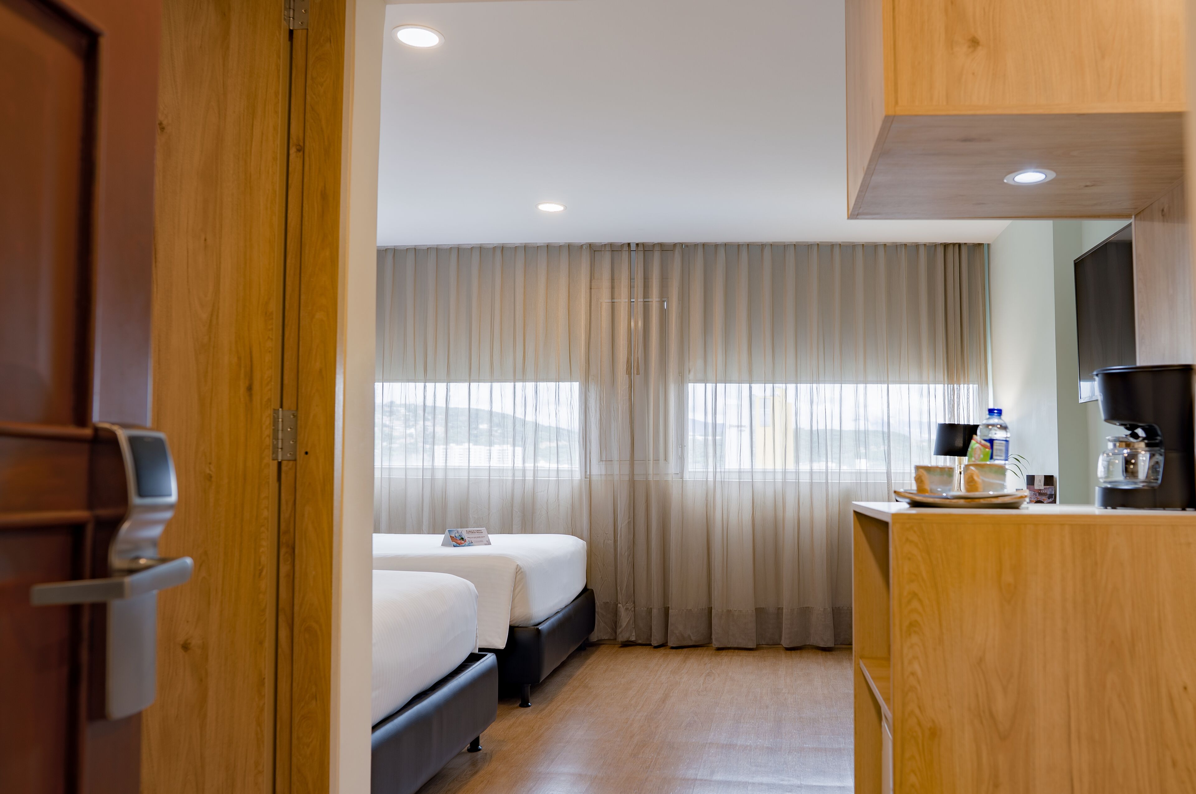 A hotel room with two beds, a wooden desk with a coffee maker, and a view through sheer curtains.
