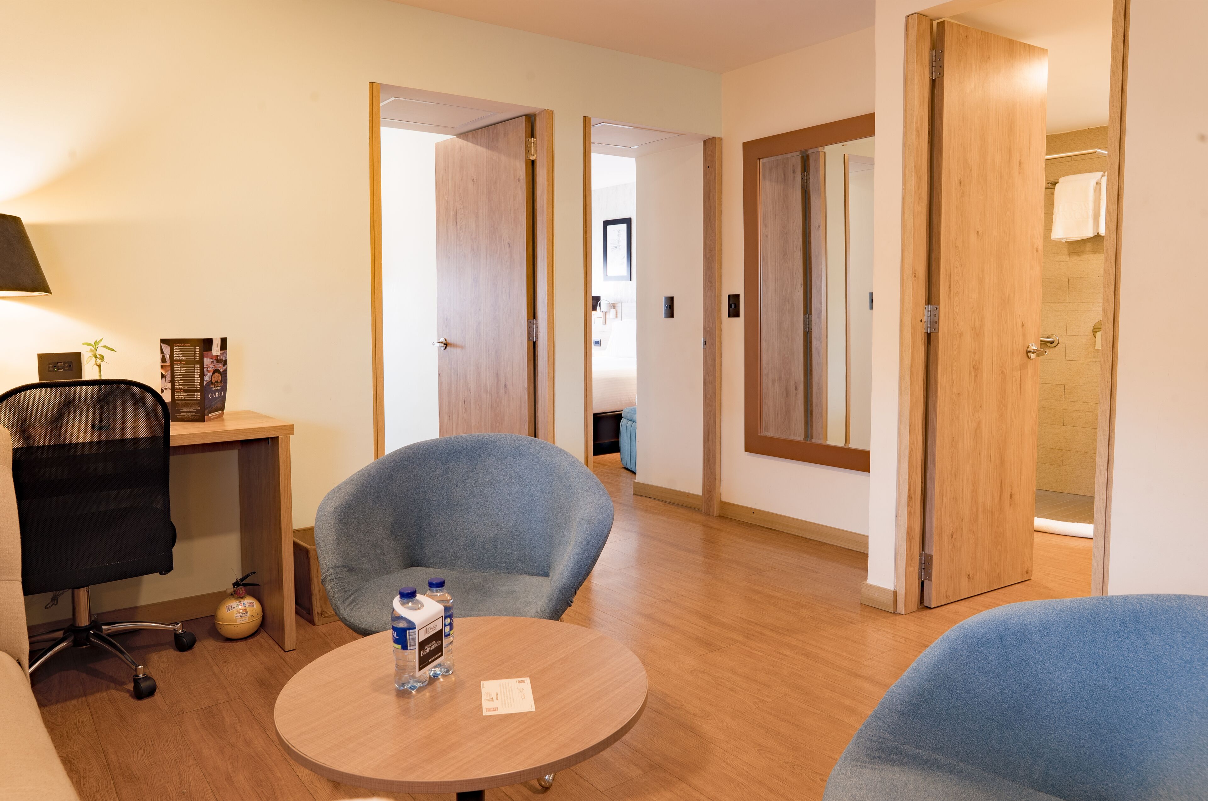 Habitación de hotel con una mesa redonda, dos sillas, un escritorio con una silla y una lámpara.