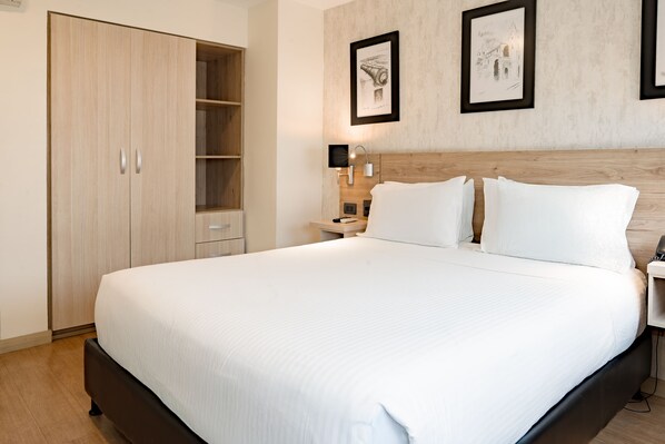 Habitación de hotel ordenada con una cama grande, un armario, una mesita de noche con lámpara y dos cuadros en la pared.