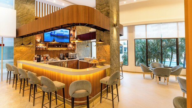 A bar area with a curved wooden counter, bar stools, a television, and a view of palm trees through large windows.