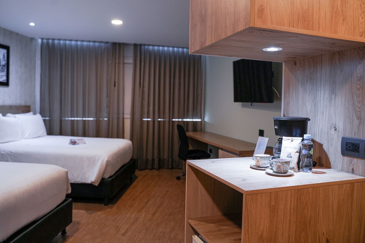 Habitación de hotel con dos camas, un escritorio con cafetera, una silla y un televisor montado en la pared.