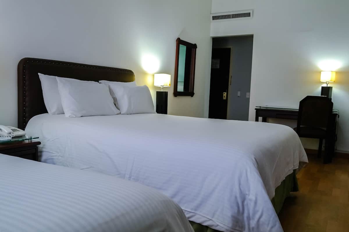 Quarto de hotel com duas camas, luminárias de cabeceira, uma escrivaninha e um telefone.