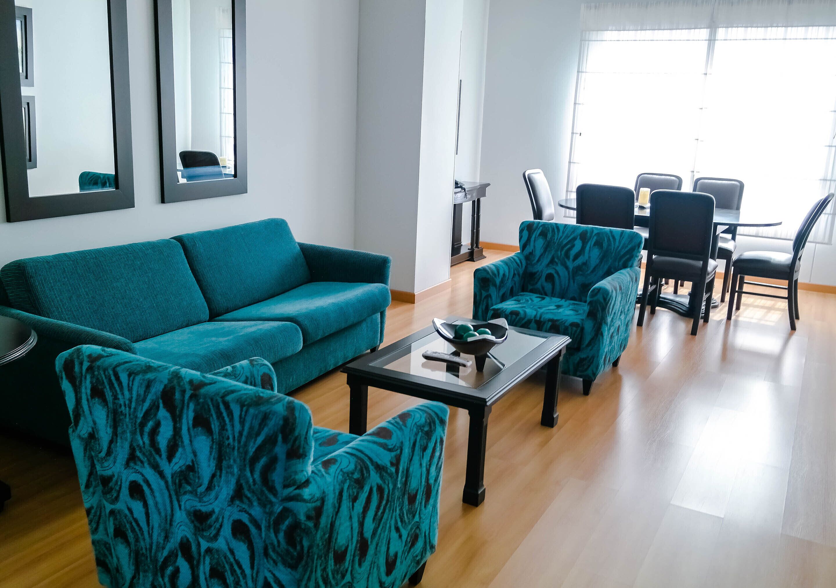 A living room with teal furniture, a glass coffee table, and a dining area with chairs and a table.