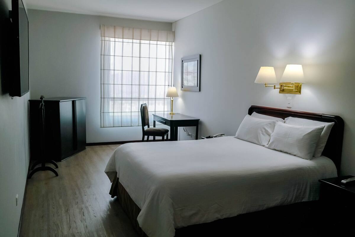 Quarto de hotel com uma cama grande, uma escrivaninha com cadeira, uma televisão e duas luminárias fixadas na parede.