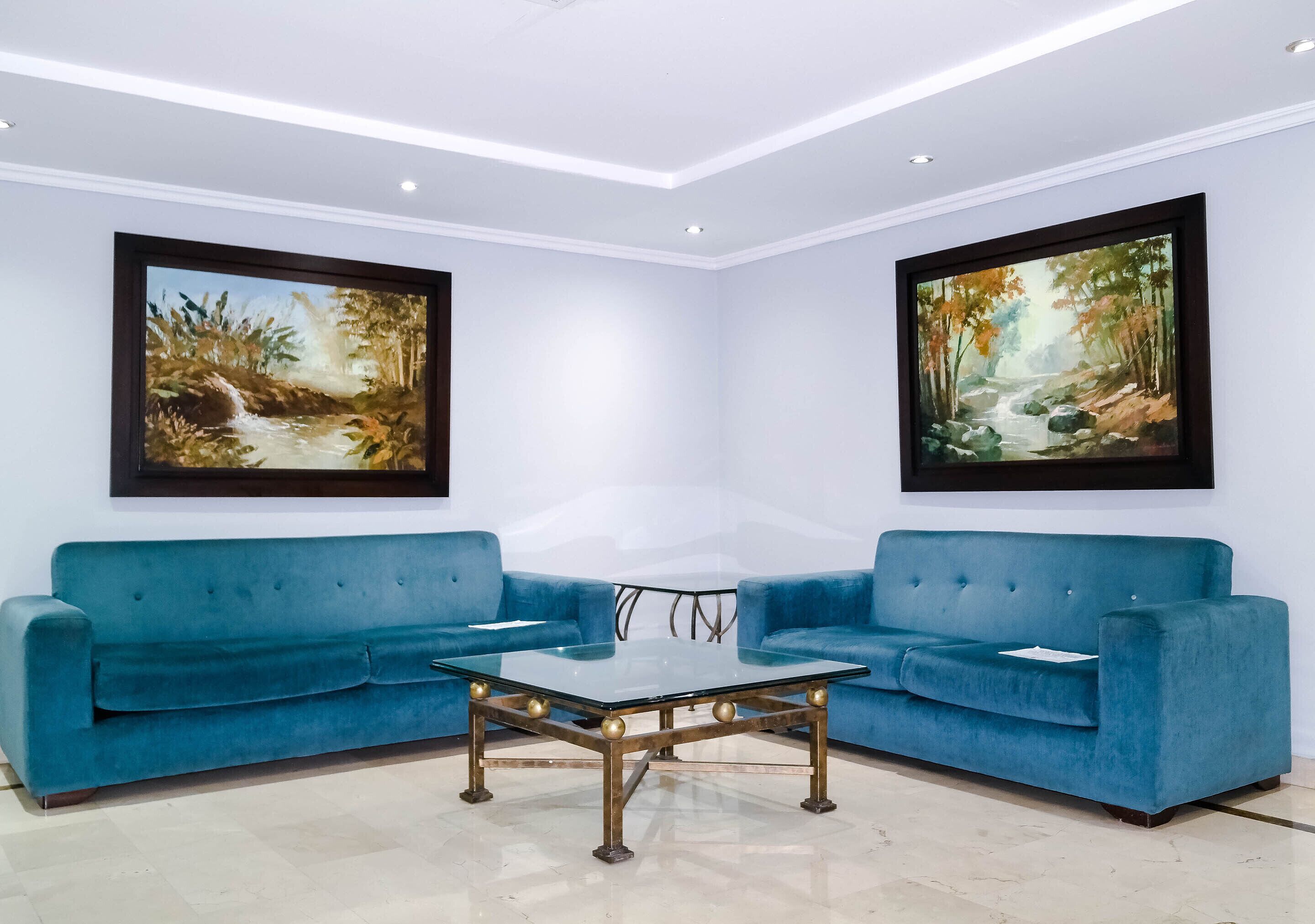 Una habitación con un sofá azul, una mesa de centro de vidrio y dos cuadros en las paredes.