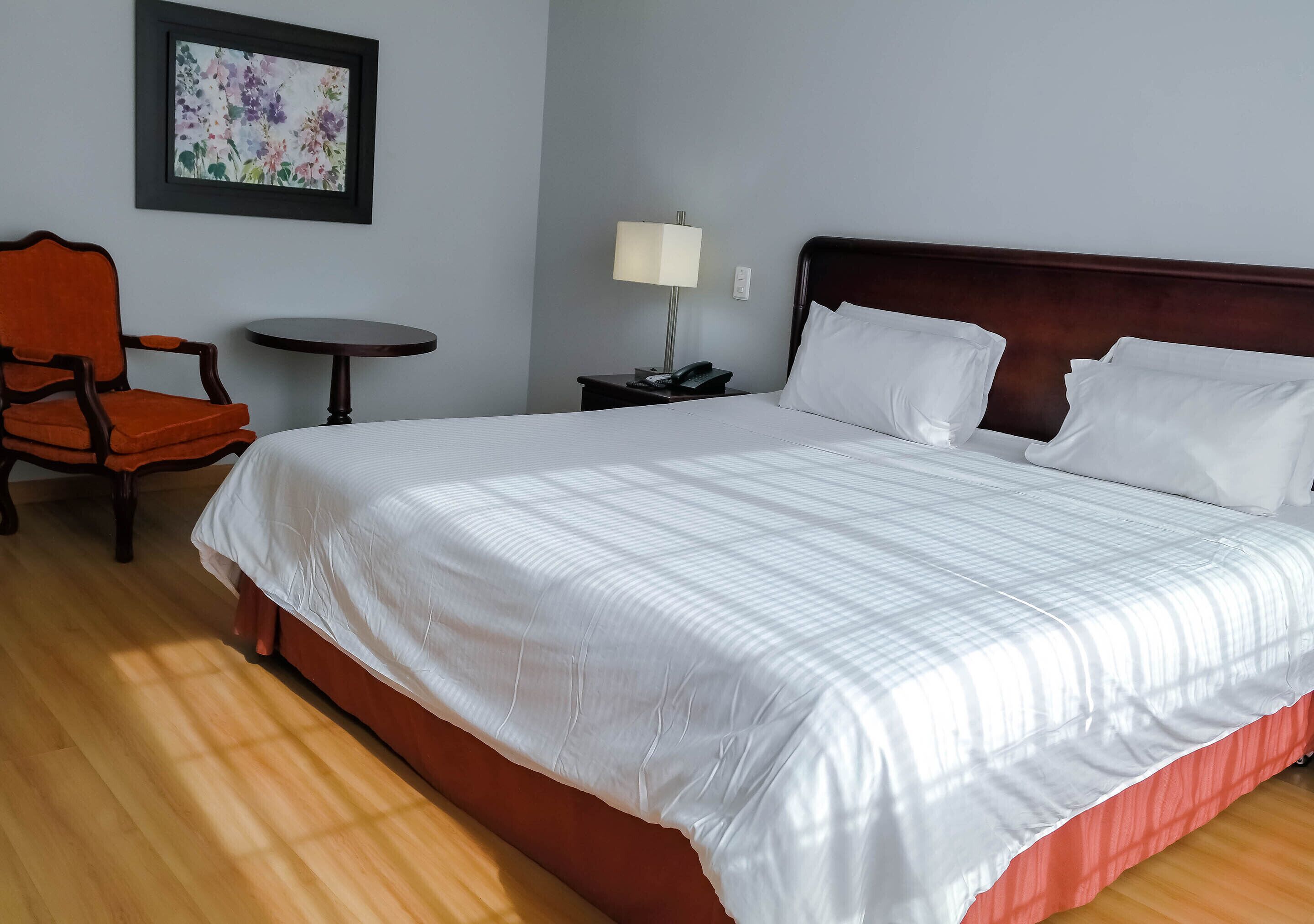 A hotel room with a large bed, a chair, a small table, and a nightstand with a lamp.