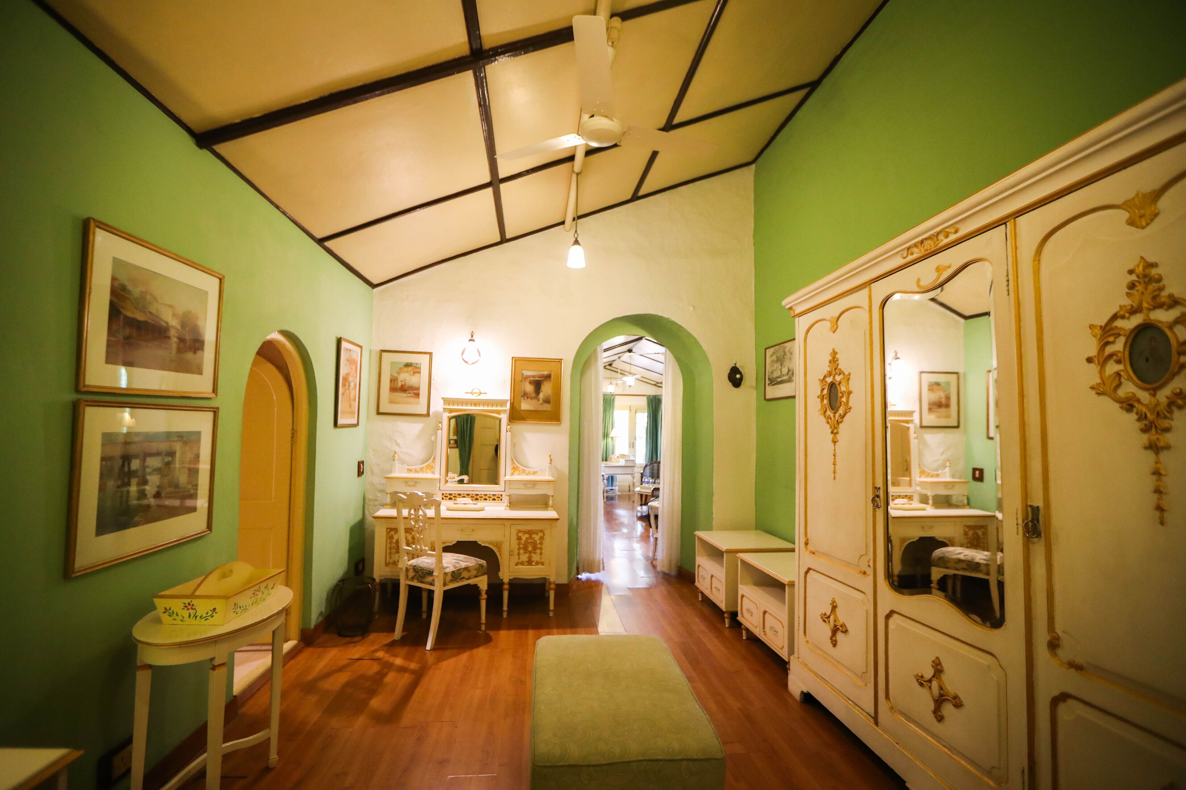 Une pièce dotée d’un plancher en bois, de murs verts, d’un plafond blanc avec des poutres noires, d’un banc vert, d’une coiffeuse blanche avec un miroir et d’une armoire blanche ornée de motifs dorés.