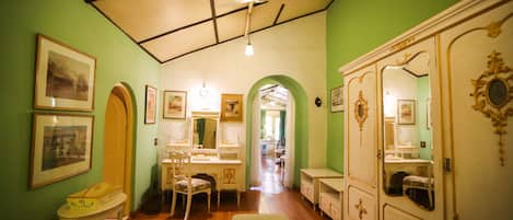 Une pièce dotée d’un plancher en bois, de murs verts, d’un plafond blanc avec des poutres noires, d’un banc vert, d’une coiffeuse blanche avec un miroir et d’une armoire blanche ornée de motifs dorés.