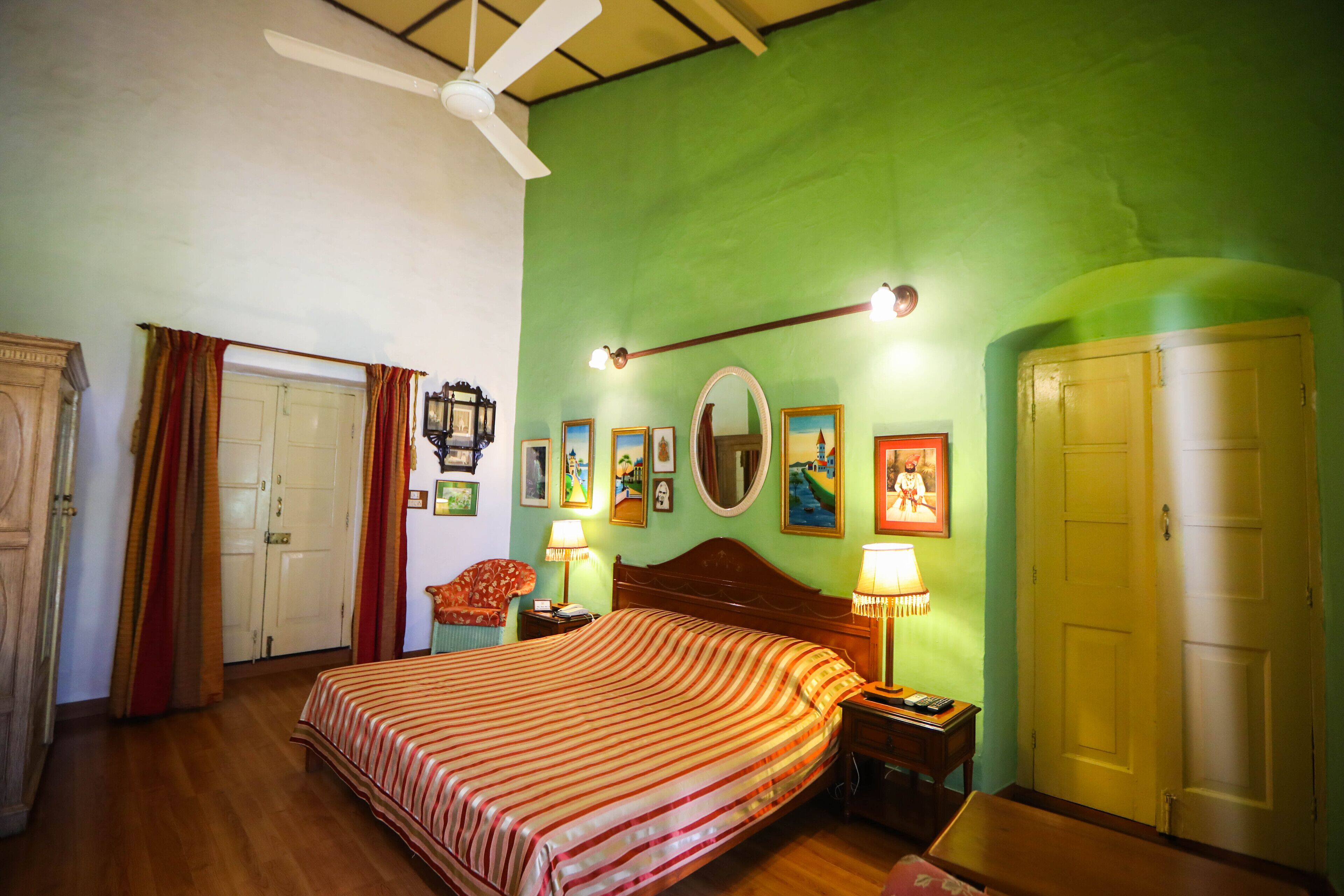 Habitación de hotel con una cama, un ventilador de techo, una mesita de noche de madera, una lámpara, un espejo y una puerta.