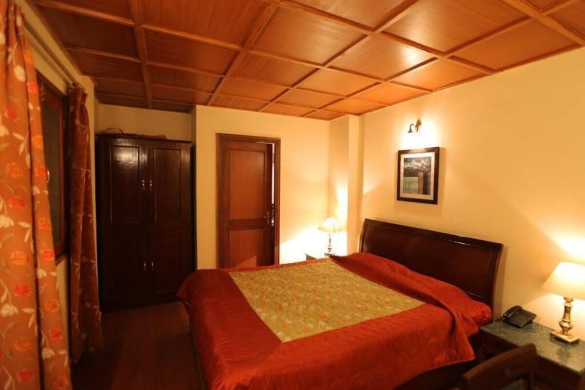 A hotel room with a wooden ceiling, a bed with a red bedspread, two bedside lamps, a wooden wardrobe, and a framed picture on the wall.