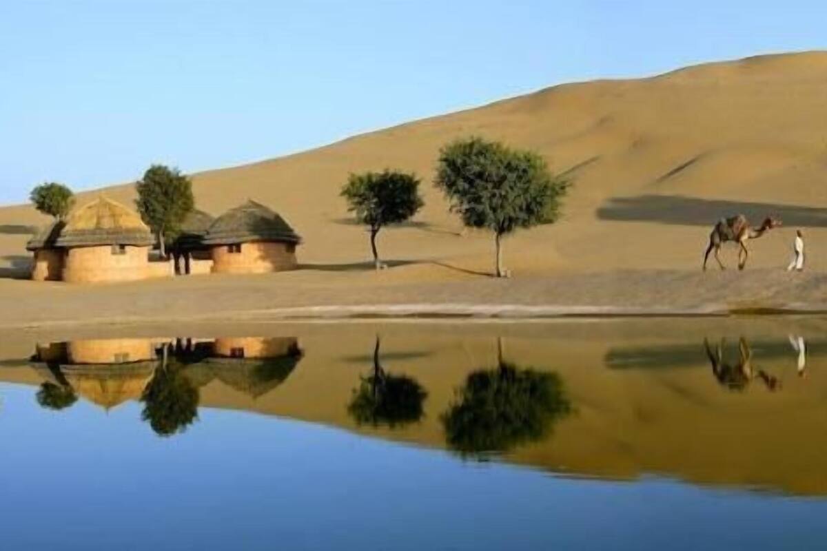 A desert landscape with sand dunes, a body of water reflecting the scene, and a few trees.