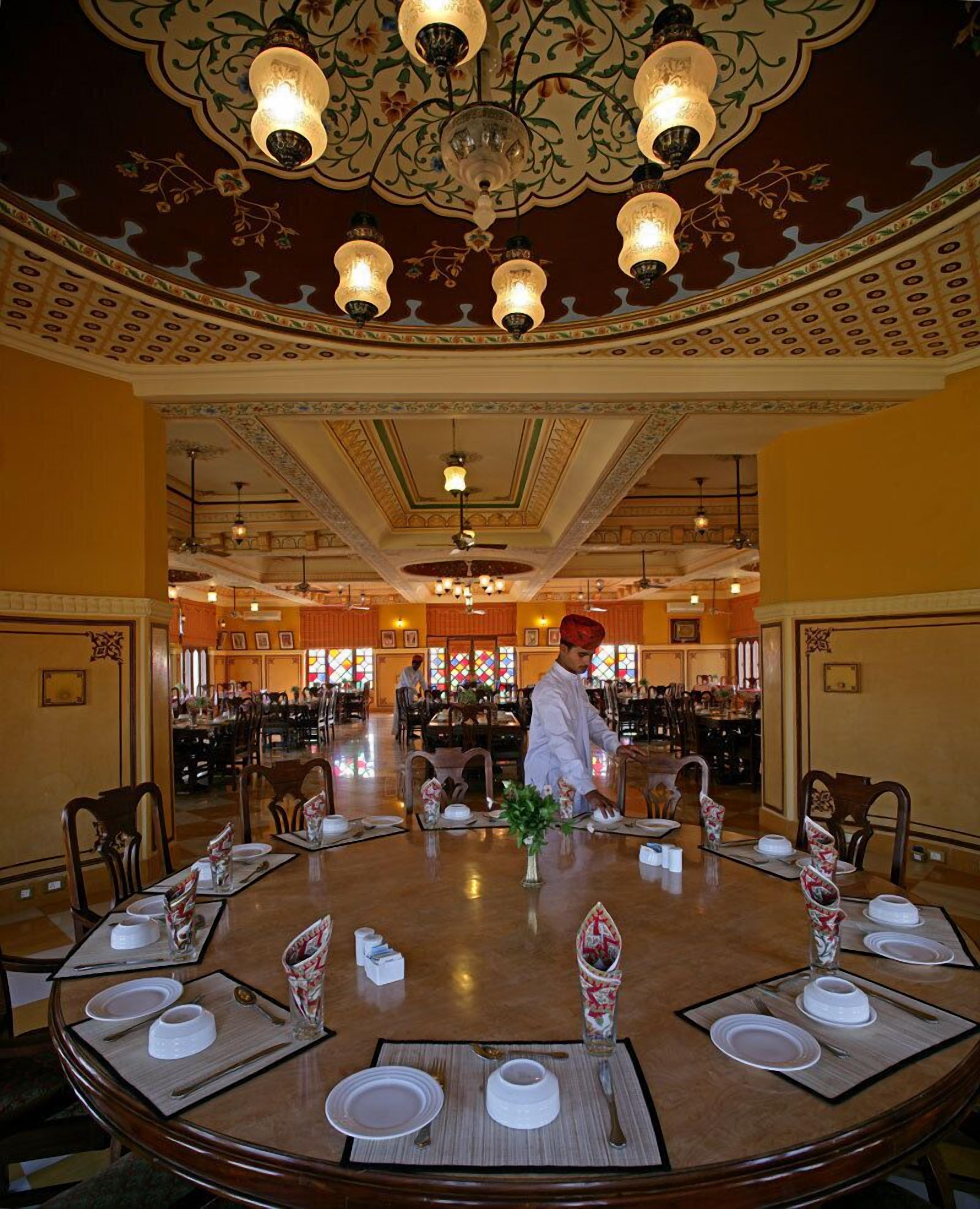 A dining area with round tables set for a meal, a chef preparing food, and a decorative ceiling with chandeliers.