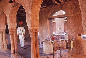 Un edificio histórico con columnas de piedra arqueadas, una mesa central con mantel decorativo, y una persona vestida con traje tradicional caminando por el lugar.