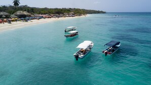 Una playa con aguas turquesas cristalinas, varias embarcaciones ancladas y una zona de arena con sombrillas y construcciones de techo de paja.