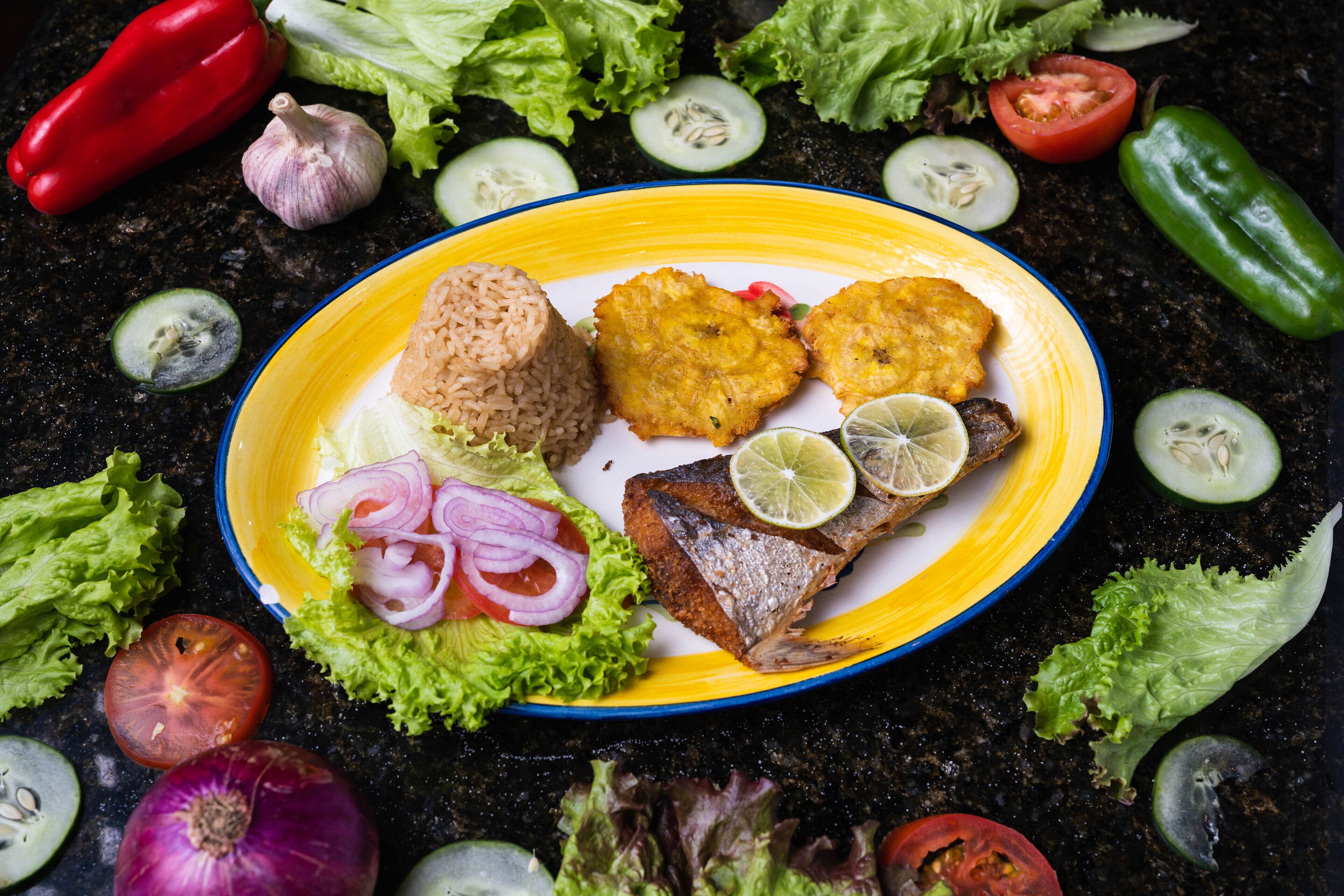 A plate of food with rice, fried plantains, fish, and lime slices, surrounded by various vegetables.