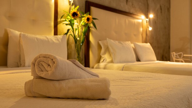 Una cama bien hecha con una toalla enrollada, un jarrón con girasoles y una luz nocturna en la pared.