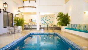Una piscina con mosaicos azules, rodeada de paredes blancas, una zona de estar con sofá y una zona de comedor con sillas y mesas.
