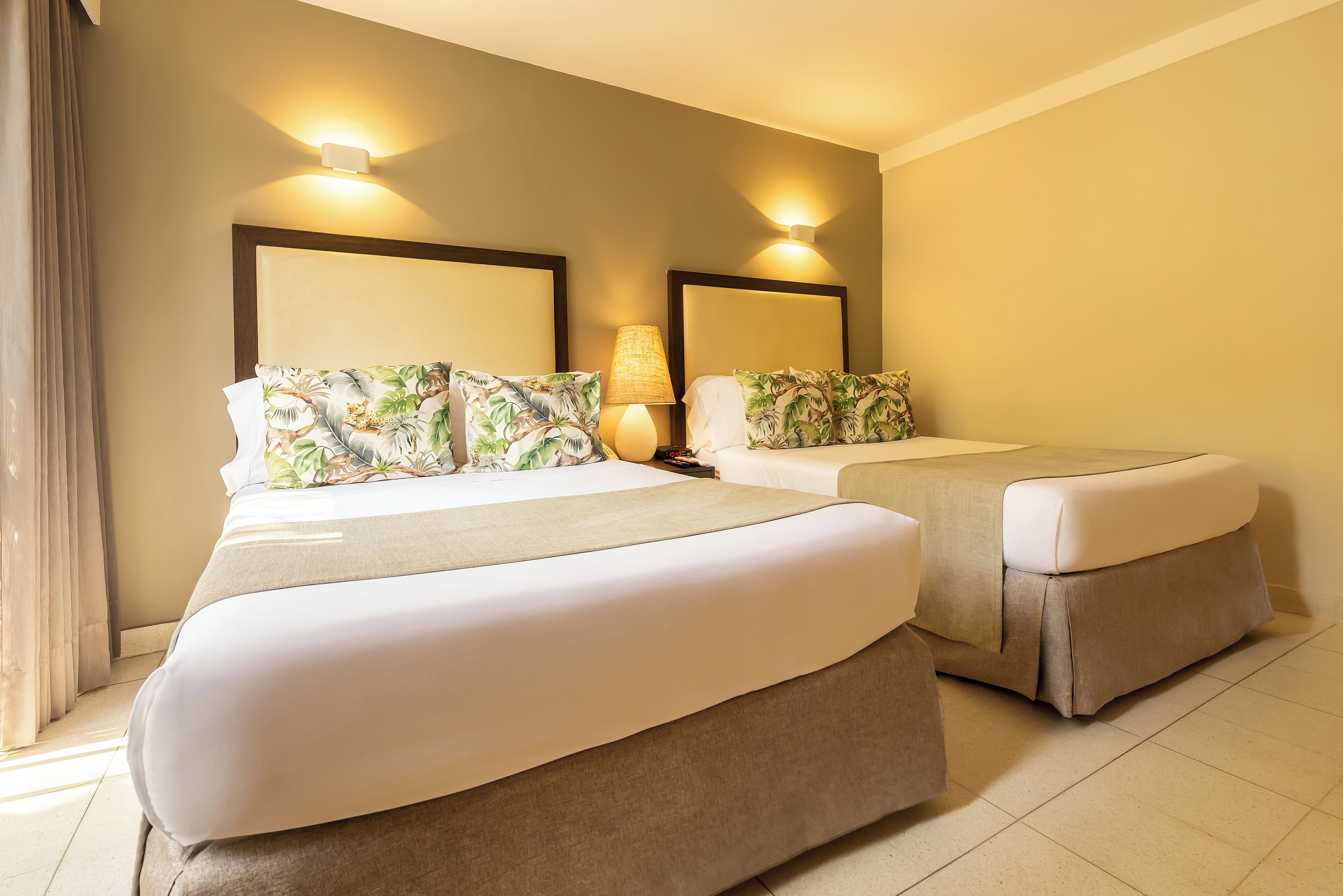 A hotel room with two beds, each with a headboard and matching pillows. There is a lamp on one side of the room.