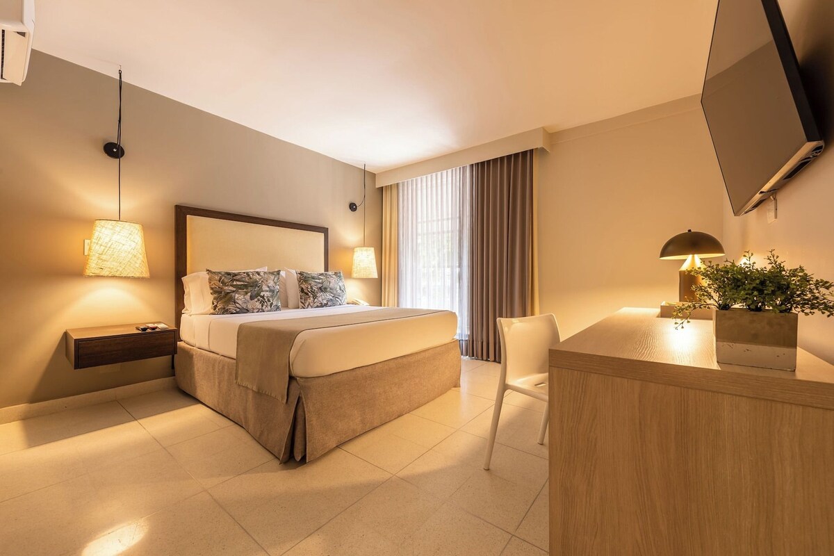 A hotel room with a bed, bedside tables, a desk, a chair, a TV, and a window with curtains.