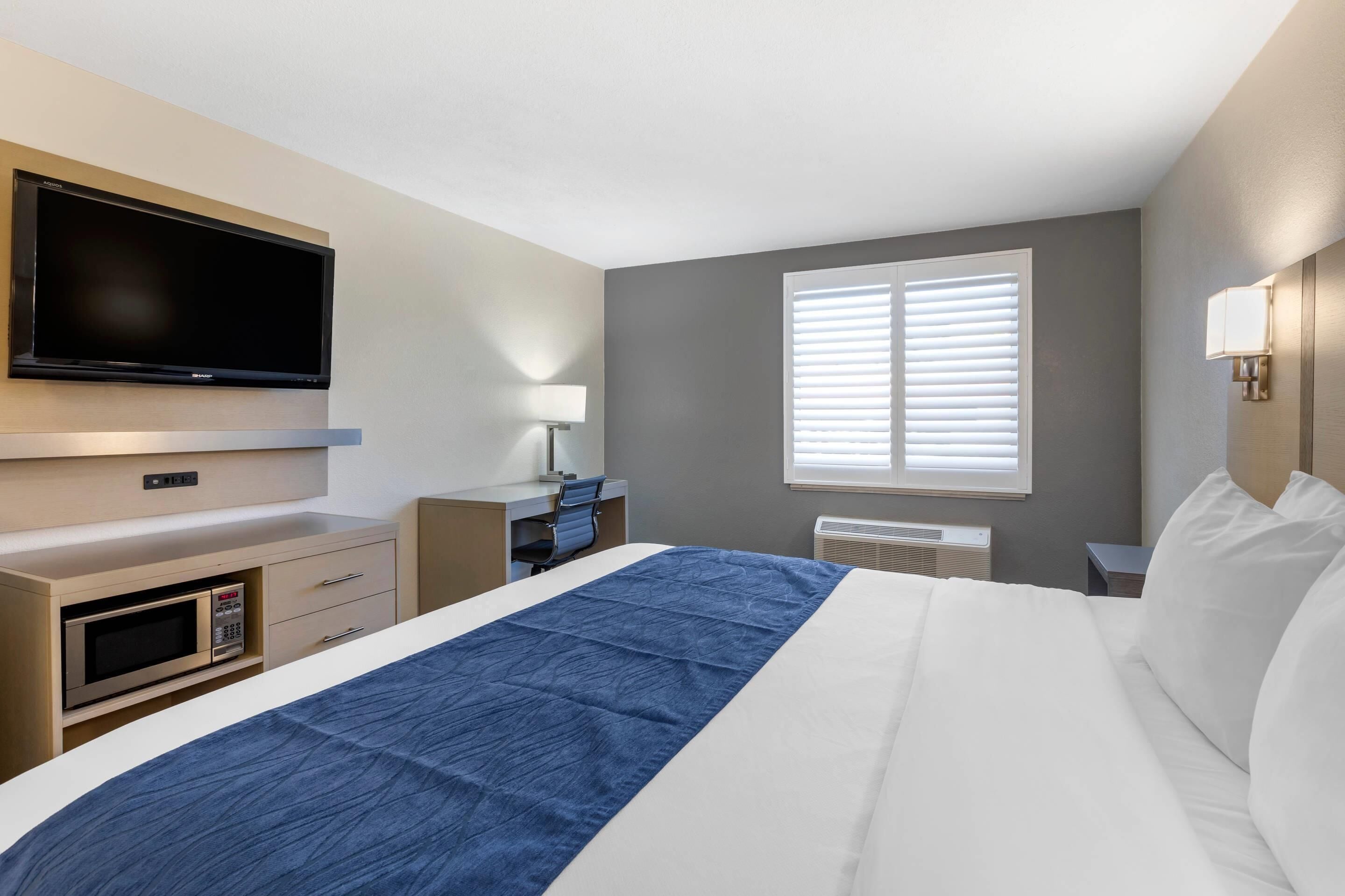 Ein Hotelzimmer mit Bett, Schreibtisch, Flachbildfernseher, Mikrowelle, Lampe und einem Fenster mit Jalousien.