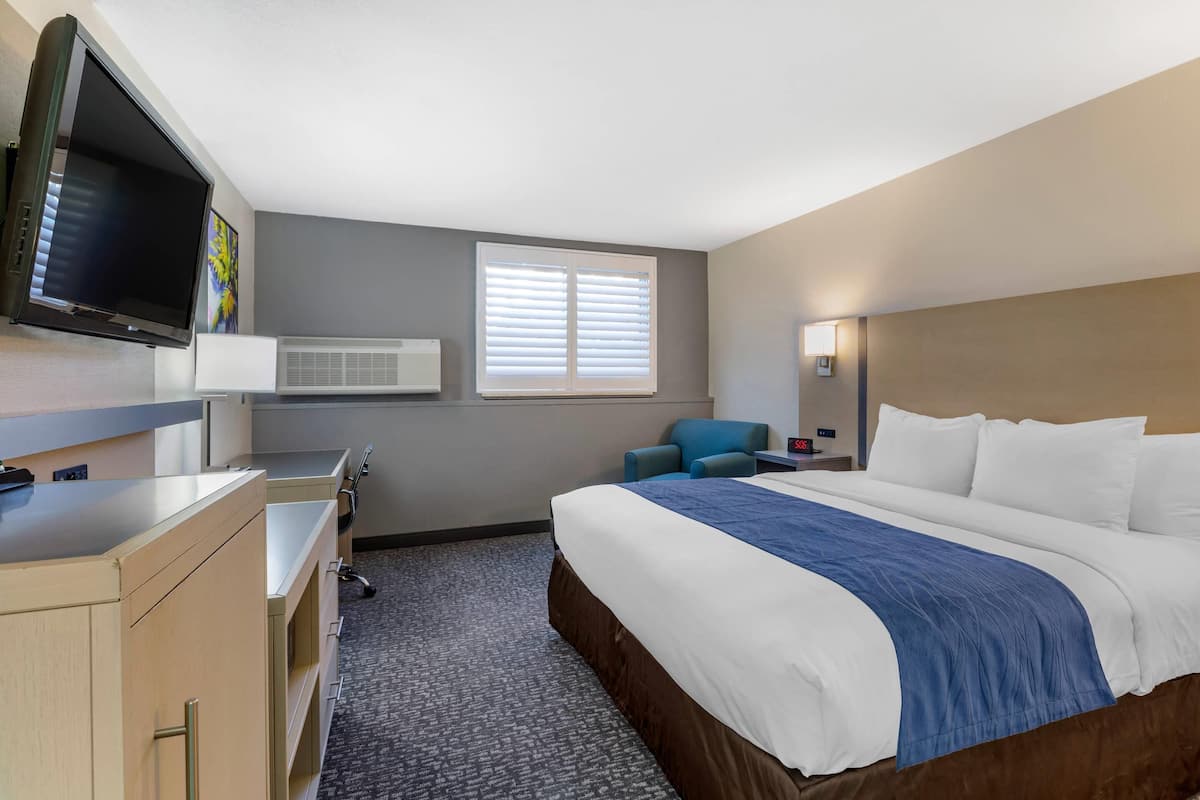 A hotel room with a large bed, a desk with a computer, a chair, a TV, and a window with blinds.