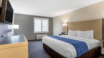 A hotel room with a bed, a desk, a flat-screen TV, a window with blinds, and a nightstand with a lamp.