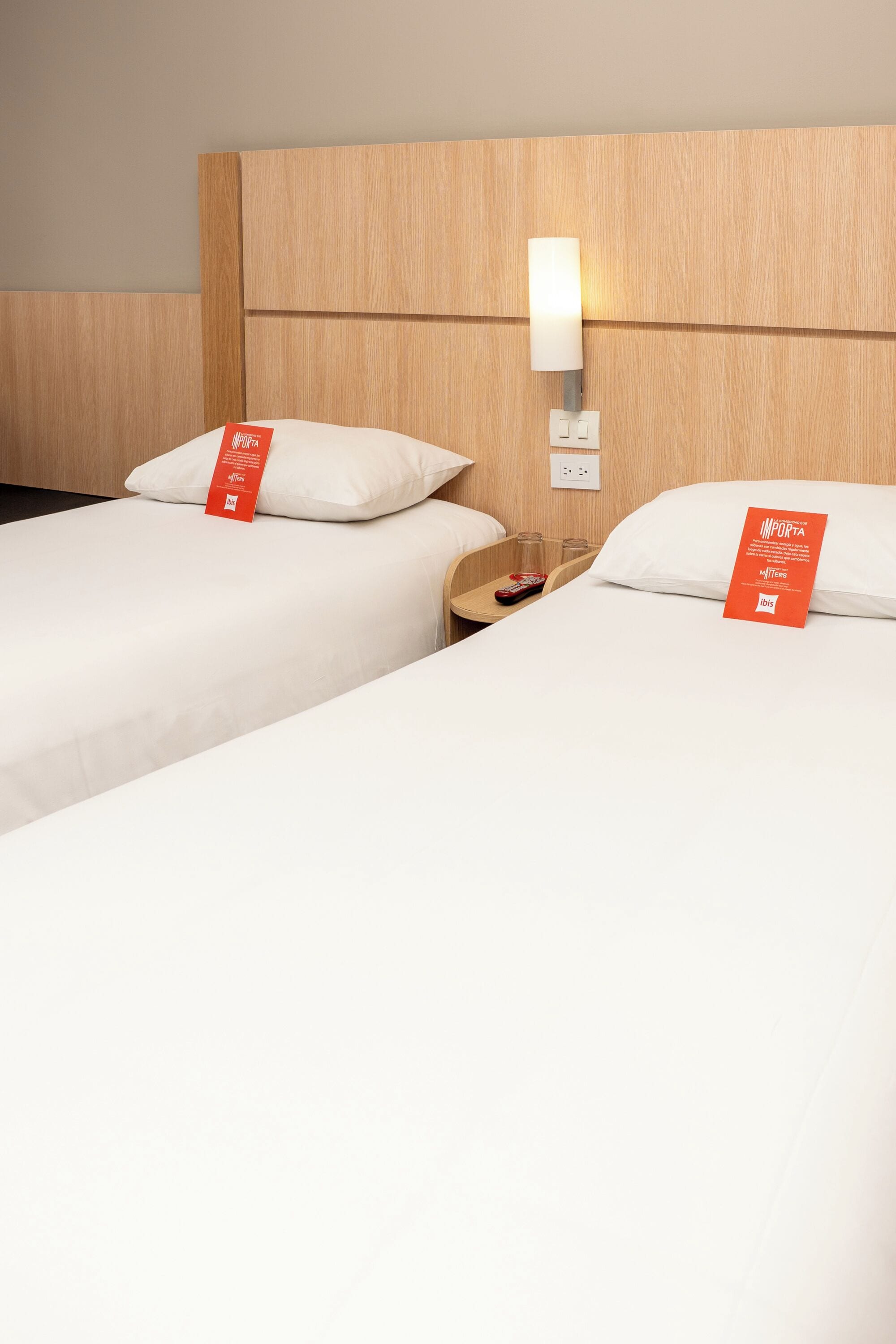 Une chambre d’hôtel avec deux lits, une table de chevet, une lampe et une étiquette rouge sur les lits.