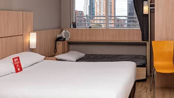 A modern hotel room with a large bed, a desk, a chair, a window with a city view, and a built-in heater.