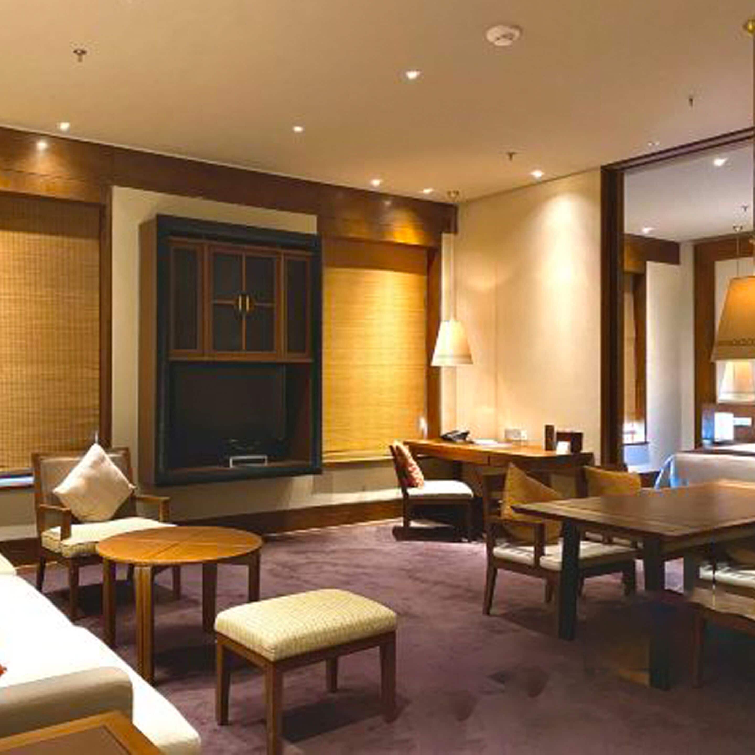 Una habitación de hotel con una cama grande, una zona de estar con una mesa y sillas, televisión y una ventana con persianas.
