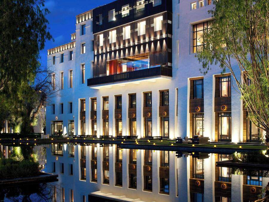 Un hotel moderno con una piscina reflectante al frente, que presenta una estructura de varios pisos con balcones y ventanas grandes.