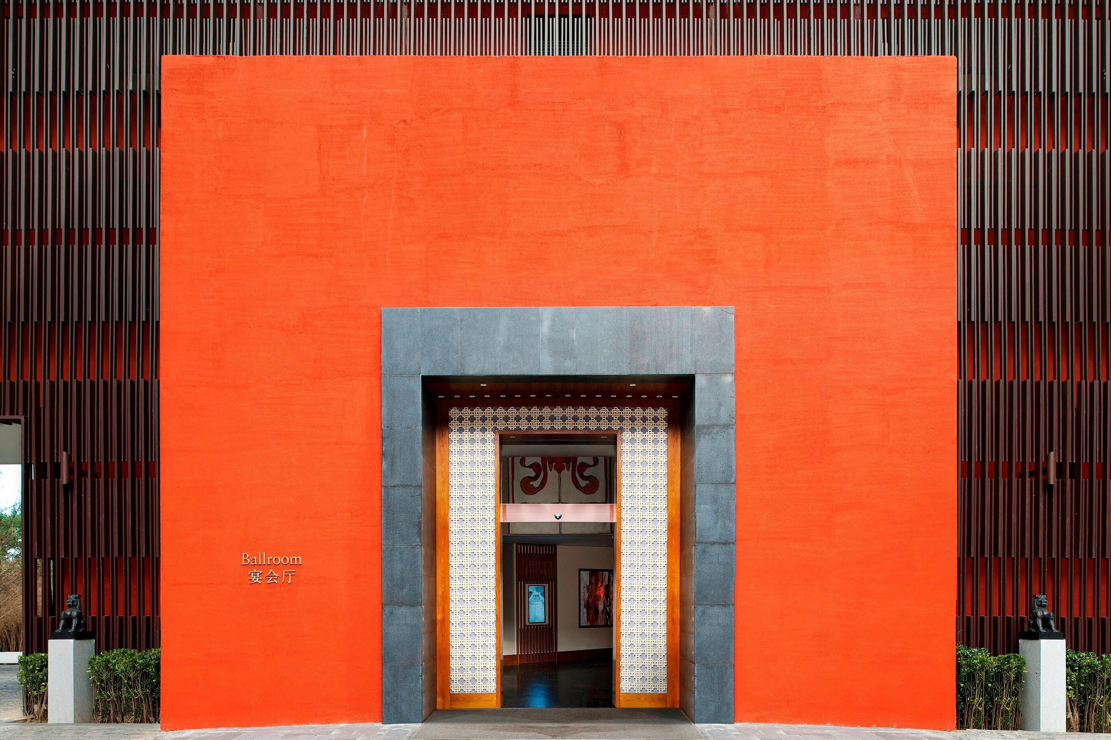 A building entrance with a large orange wall, a framed doorway, and a small statue outside.