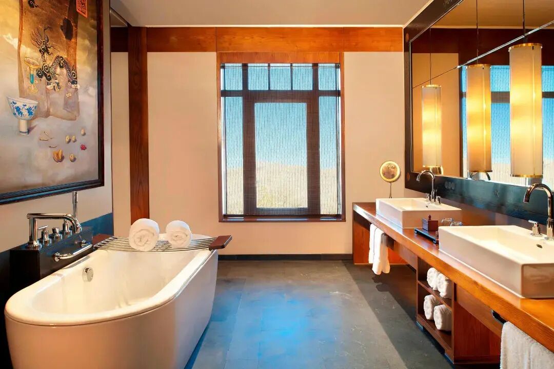 A modern bathroom with a freestanding bathtub, two sinks, a large mirror, and a window with vertical blinds.