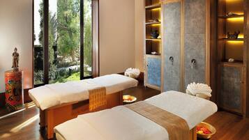 A spa room with two massage tables, a large cabinet with shelves, and a view of greenery outside.
